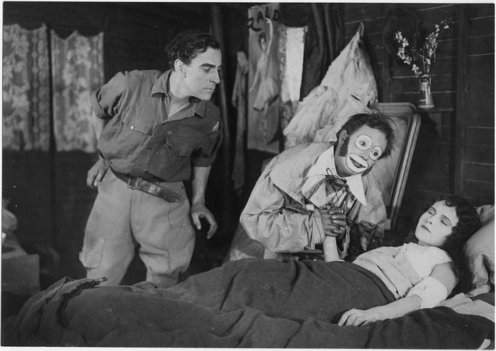 Une jeune femme alitée visitée par un clown et un homme se penchant vers le lit