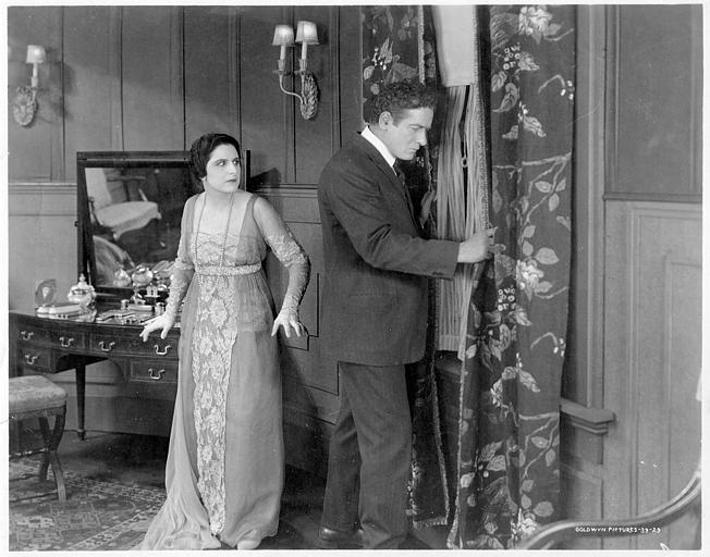 Géraldine Farrar est aux côtés d'un homme qui espionne à la fenêtre derrière un rideau