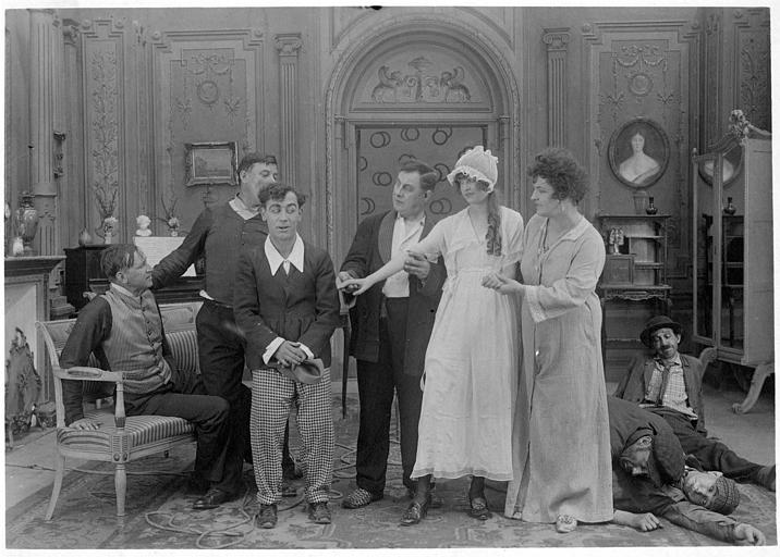 Dans un salon, Rémond Frau dans le rôle de Dandy, est timide devant Pansy Wilde qu'on lui présente entourée de et plusieurs personnages Madame Devries, Smote, Riche, Gaston, Max, Brenie et Gohier dans 'Dandy-afficheur' de Georges Rémond