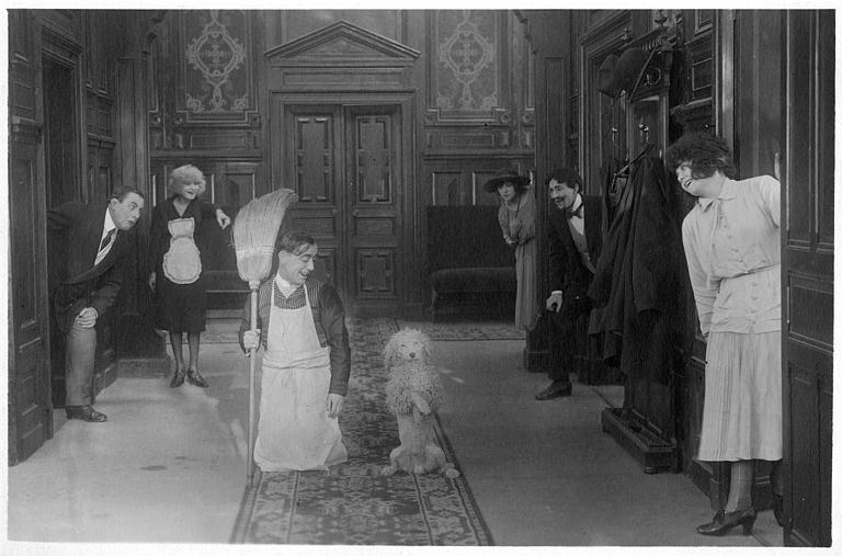 Dans un couloir, Rémond Frau dans le rôle de Dandy à genoux à côté d'un chien dressé tandis que Ulysse, Florelle, Max et d'autres l'encouragent sur le pas de chaque porte