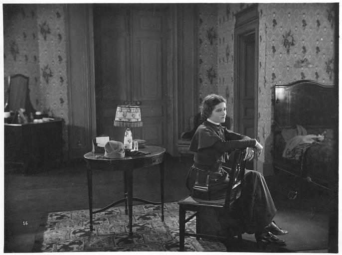 Marie Glory (alias Arlette Genny) dans le rôle de Miss Helyett assise les bras appuyés sur le dossier de sa chaise dans une chambre