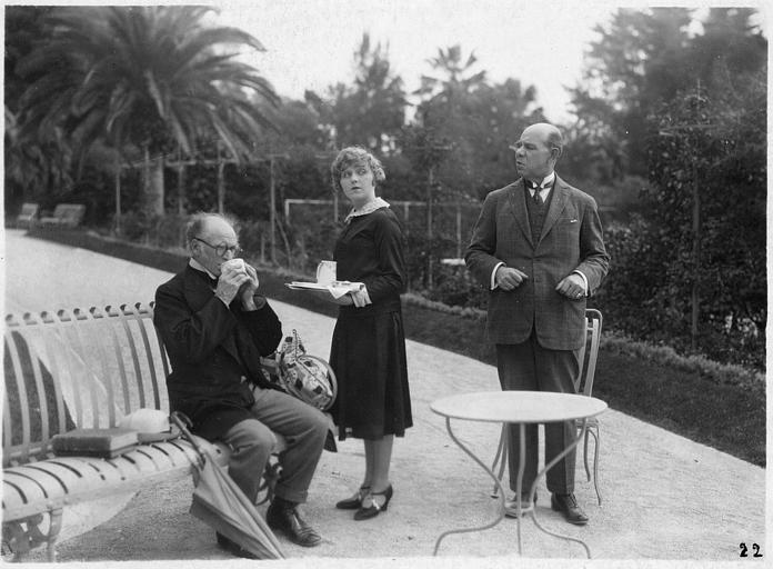 Fred Wright est assis sur un banc pour boire le verre que lui a apporté Betty Balfour, tandis que Louis Baron le regarde