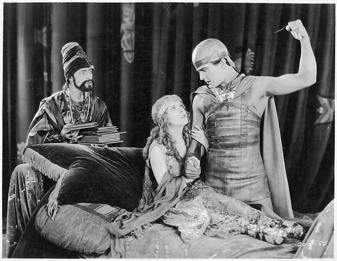 Une homme levant son poing devant May McAvoy assise sur un lit