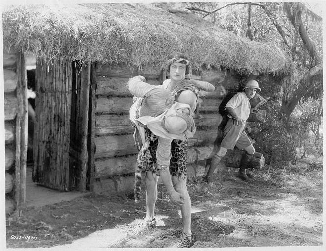 Franck Merril dans le rôle de Tarzan portant un homme à l'entrée d'une cabane tandis qu'un autre s'enfuit
