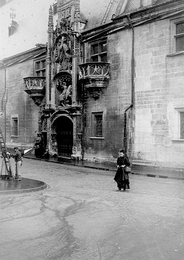 Vue du cartouche de l'entrée devant lequel passe une femme, un homme installe un drapeau à l'aide d'une échelle