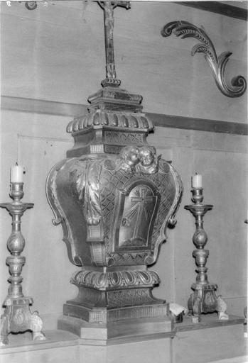 Tabernacle en forme d'urne à la porte ornée d'une croix, décor de chérubins, volutes, feuilles d'acanthe et oves, surmonté d'une croix d'autel, bois taillé, doré