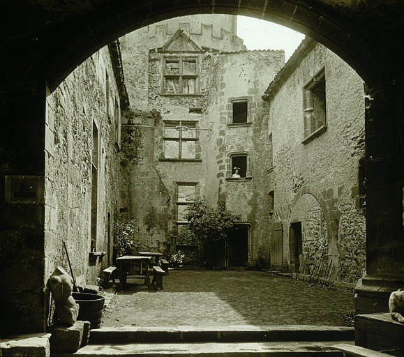 Vue des façades intérieures sur cour avec un passage voûté au premier plan et une femme portant chapeau dans l'encadrement d'une fenêtre