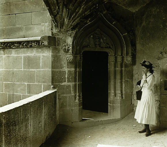 Vue prise à l'intérieur du château, porte ouverte sur une tourelle d'escalier, une jeune femme debout en retrait de l'ouverture de la galerie-haute avec un appareil photographique à la main et son étui en bandoulière