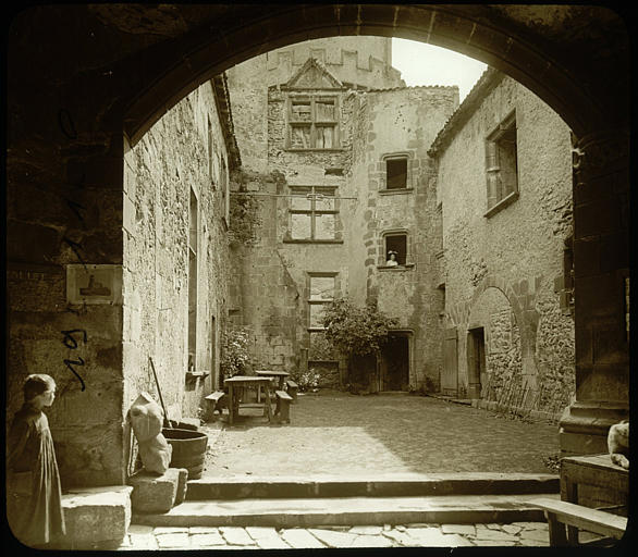 Vue de façades intérieures sur cour avec un passage voûté au premier plan et une femme portant chapeau dans l'encadrement d'une fenêtre, une petite fille à gauche