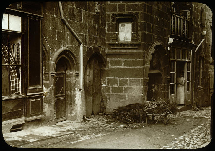 Façade sur la rue, partie basse, du bois est déposé devant la porte de la maison, un homme se tient sur la porte d'une boutique