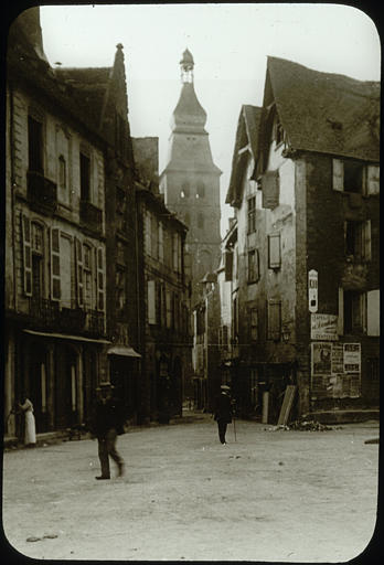 Place et des façades de maisons, à l'arrière-plan le clocher de la cathédrale, personnages