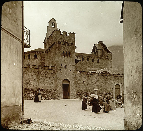 Vue d'ensemble depuis la place située devant l'église, portail, enceinte, nef et clocher à l'arrière-plan, scène de marché avec des personnages