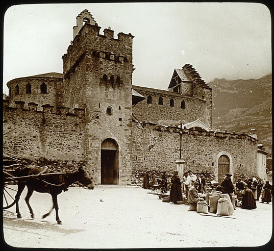 Vue d'ensemble depuis la place située devant l'église, portail, enceinte, nef et clocher à l'arrière-plan, scène de marché avec des personnages, un âne