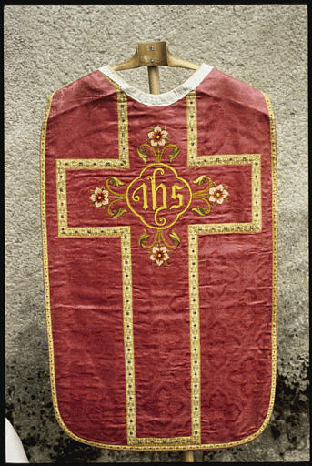 Chasuble : tissu damassé rouge à motifs quadrilobés et motifs végétaux, croix centrale formée par un galon doré, fleurs et inscription brodées