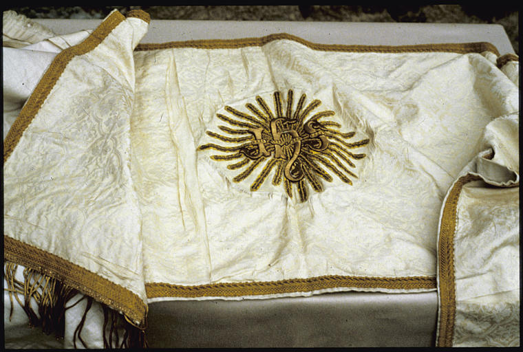 Dais de procession : tissu damassé blanc, motifs quadrilobés, feuillages, galon doré, au centre, inscription entourée de rayons brodés