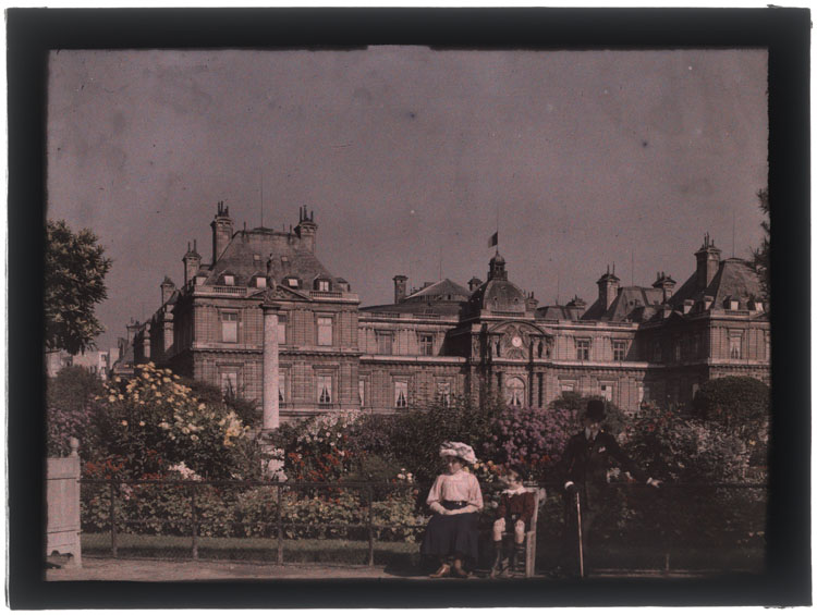 Marthe Antoine et un petit garçon assis devant un parterre de fleurs, Jean Antoine portant chapeau melon et canne debout à leurs côtés, le palais en arrière-plan