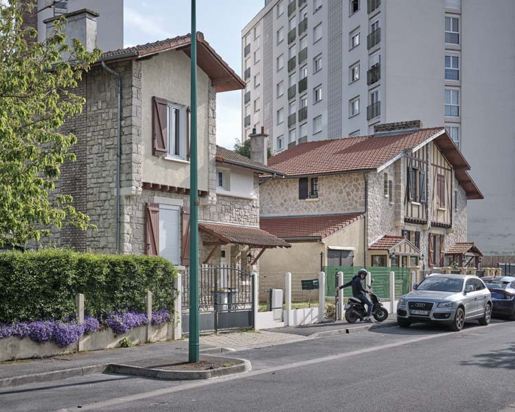 Depuis la rue, alignement de maisons jumelées ; Barres d’immeubles à l’arrière-plan