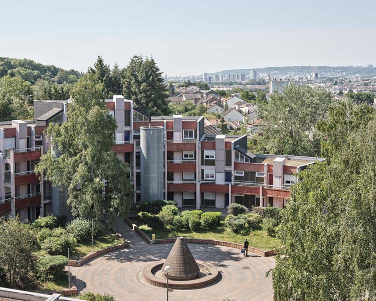 Seconde ligne de bâtiment, enroulée autour d’une place occupée en son centre par une fontaine
