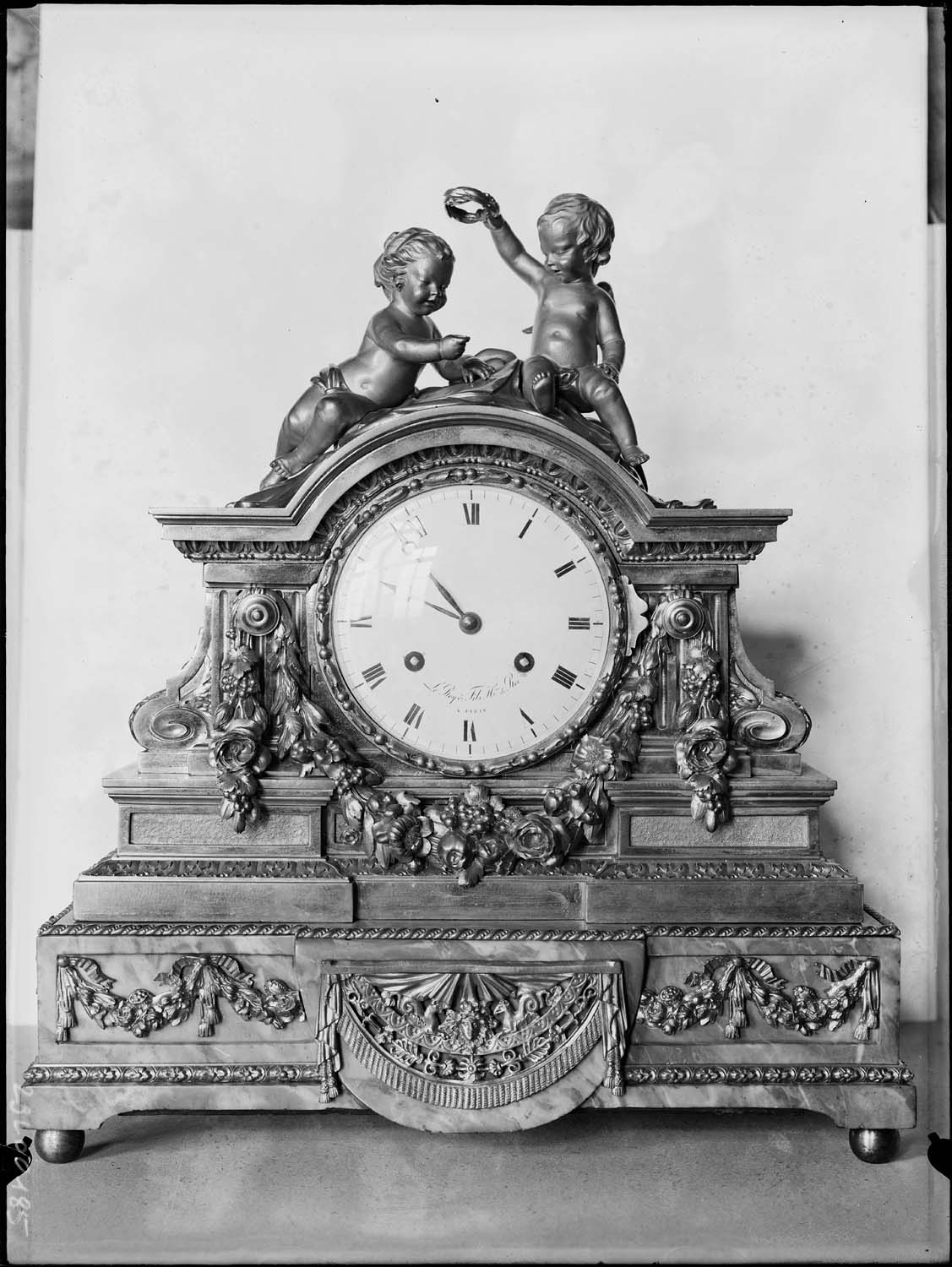 Pendule ornée de guirlandes de roses et de deux enfants (l’un posant une couronne de lauriers sur la tête de l’autre), bronze doré, époque Louis XVI, Inv. B180, classée Monuments historiques le 15 mai 1980.