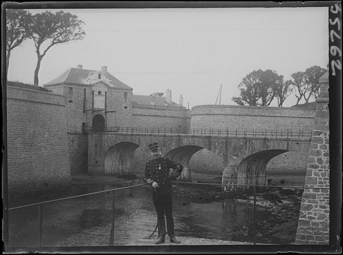 Vue d'ensemble du pont d'accès, vers le pont-levis et l'enceinte fortifiée : officier posant au premier plan