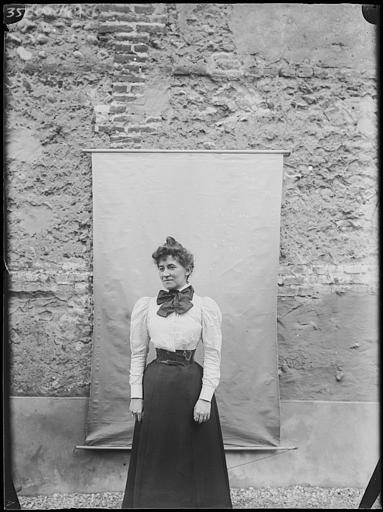 Portrait de Mme Huguet, en pose debout, de face devant un mur recouvert d'une toile suspendue