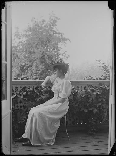 Scène de genre : jeune femme assise sur une terrasse, accoudée à une rambarde, vue au travers d'une porte-fenêtre ouverte