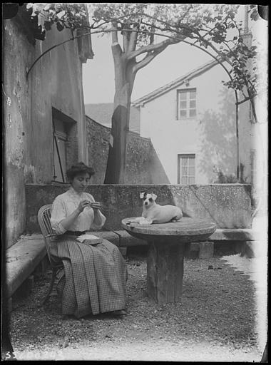Cour : femme assise enfilant une aiguille à coudre, son fox-terrier couché sur la table de pierre, treillage