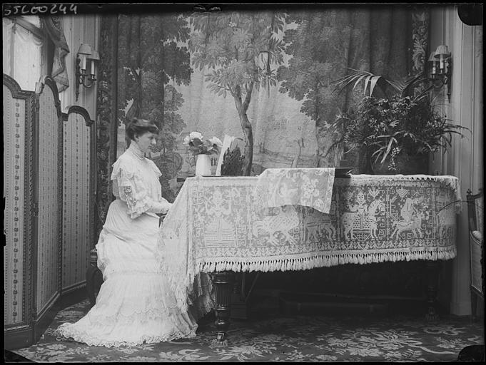 Intérieur, salon : Marcelle assise au piano, au fond tapisserie (verdure et oiseaux), parement en guipure à thème populaire pour le piano