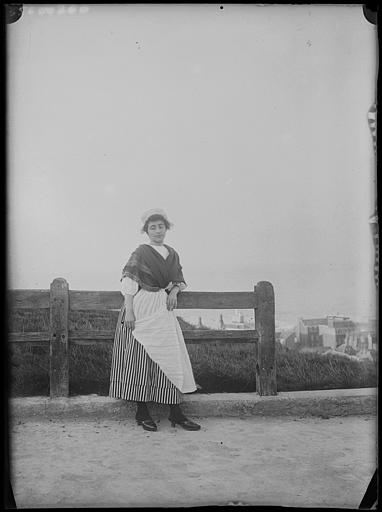 Bord de falaise, barrière de bois, vers la ville en contre-bas : jeune femme (Suzanne ou Denyse) en costume régional en pose debout