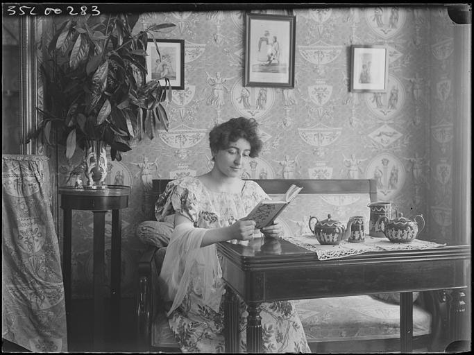 Scène de genre, intérieur, salon : jeune femme en robe de style Empire assise lisant un livre, les mains posées sur une table avec un service à thé à décor antique