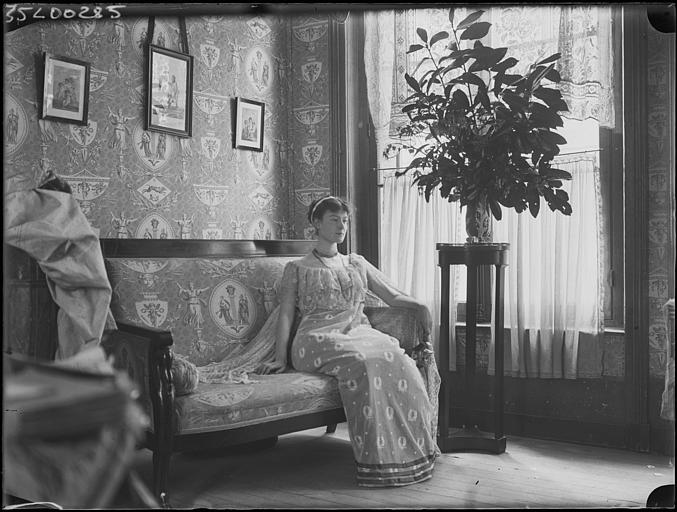 Scène de genre, intérieur, salon : jeune femme en robe de style Empire assise sur un canapé, de face, composition de feuillages sur une table haute, vers la fenêtre