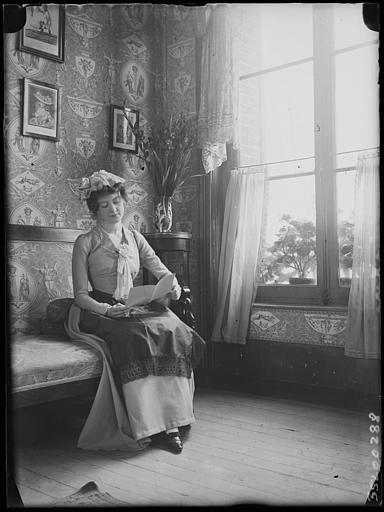 Scène de genre, intérieur, salon : jeune femme (Suzanne ou Denyse) en costume régional assise sur un canapé, de trois-quarts face, vers la fenêtre
