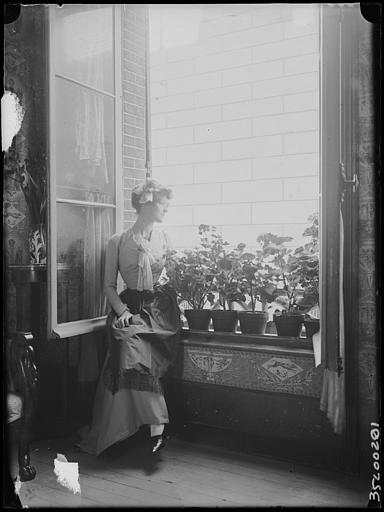 Scène de genre, intérieur, salon : jeune femme (Suzanne ou Denyse) en costume régional assise sur le rebord de la fenêtre, pots de fleurs, visage de profil