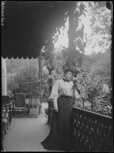 Scène de genre : femme posant sous une terrasse couverte, debout, regardant vers le jardin