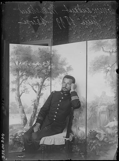 Intérieur : portrait du Commandant Marchand assis accoudé au dossier d'une chaise devant un paravent peint (scène bucolique), une main sur la cuisse