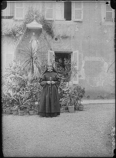 Cour : portrait d'une religieuse en pose devant la statue de la Vierge (sur la façade), série de plantes vertes derrière elle