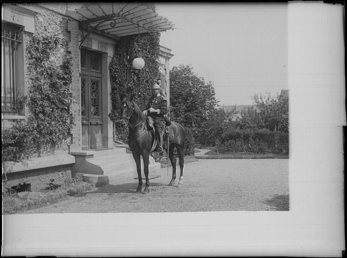 Portrait d'un officier du 18e Régiment de cavalerie, à cheval, devant la façade d'une maison