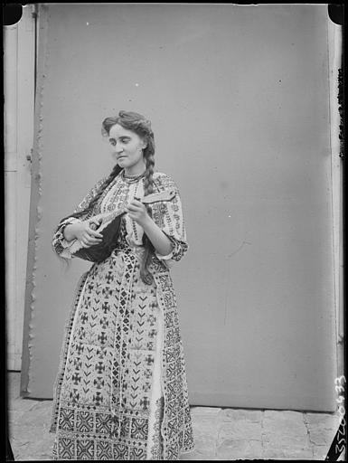 Extérieur, cour pavée, pose devant une toile : Mlle Desforges en roumaine avec une mandoline dans les mains, de trois-quarts face