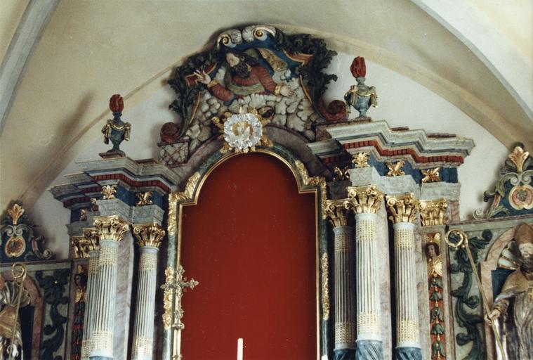 Retable : détail du fronton sculpté en bas-relief de la figure de Dieu le Père émergeant des nuées, bois sculpté, peint, doré ; après restauration