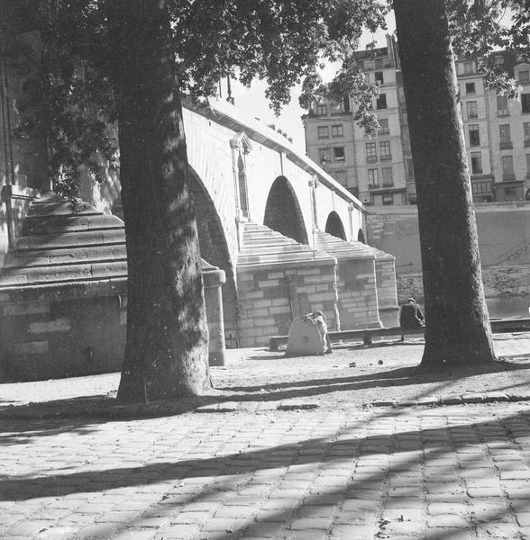 [Un homme assis sur un banc, sur un quai, au pied du pont Marie]