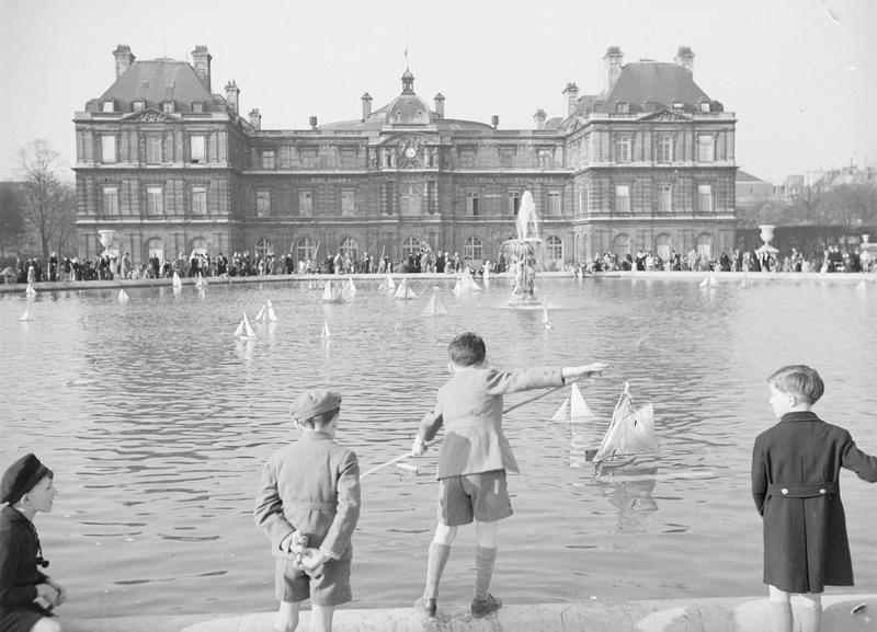 [Des enfants jouent avec des petits bateaux dans la fontaine Odéon du jardin du Luxembourg, devant le palais]