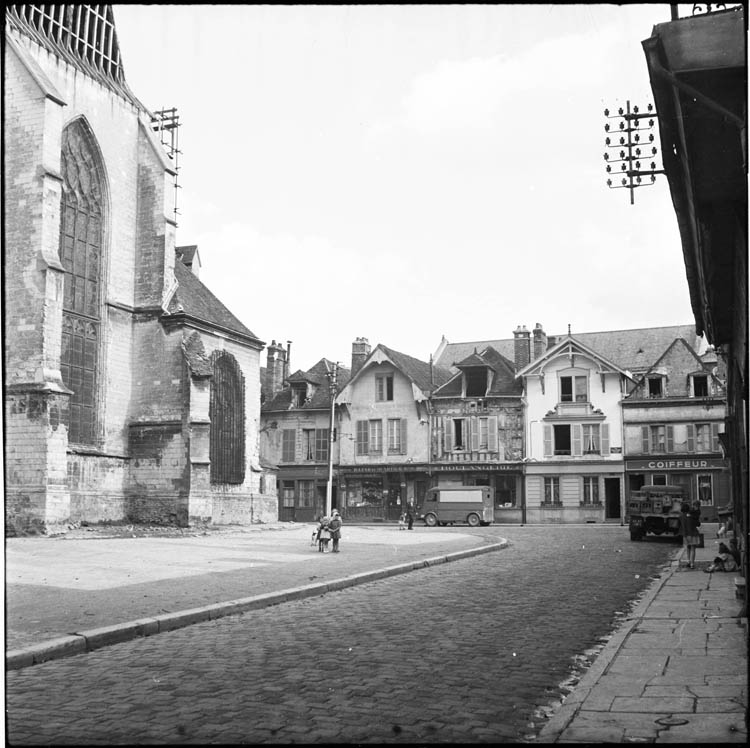 Abords ; alignement des façades sur la rue longeant l’église ; enseignes de boulangerie, de coiffeur et de « Bazar de quartier »