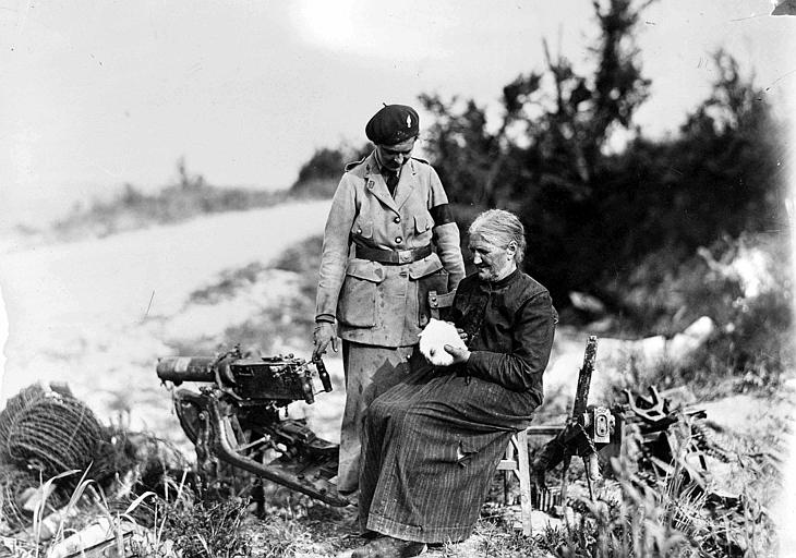 Vieille femme assise avec un chaton dans les mains et Mlle Alice Parsons, travailleuse du CARD, mitraillette sur pied et armes diverses