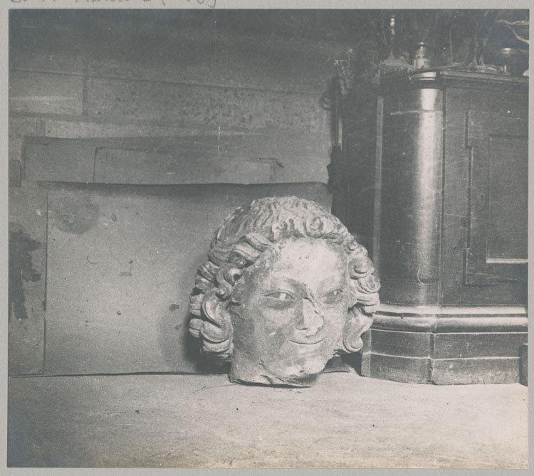 Statue : tête d'ange souriant mutilée par les bombardements, provenant du 2e contrefort de la nef, façade nord, et entreposée dans les petites sacristies