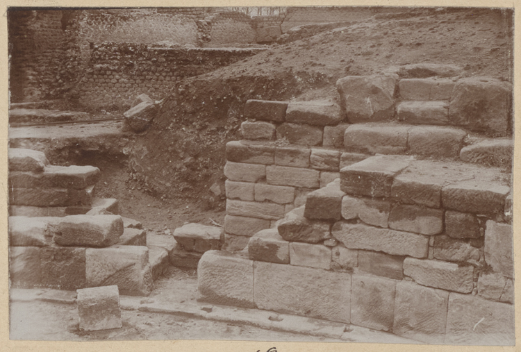 Podium (partie déblayée au nord-ouest), escaliers conduisant des gradins à l’arène et jambage de porte donnant dans un celle. Détail de cet escalier et de ce jambage de porte vus de l’arène