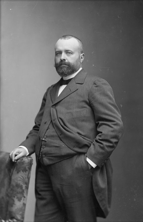 Docteur Lucien Picqué, en mi-corps, debout, pose de trois quarts, une main dans la poche de son pantalon et l'autre posée sur le dossier d'une chaise.