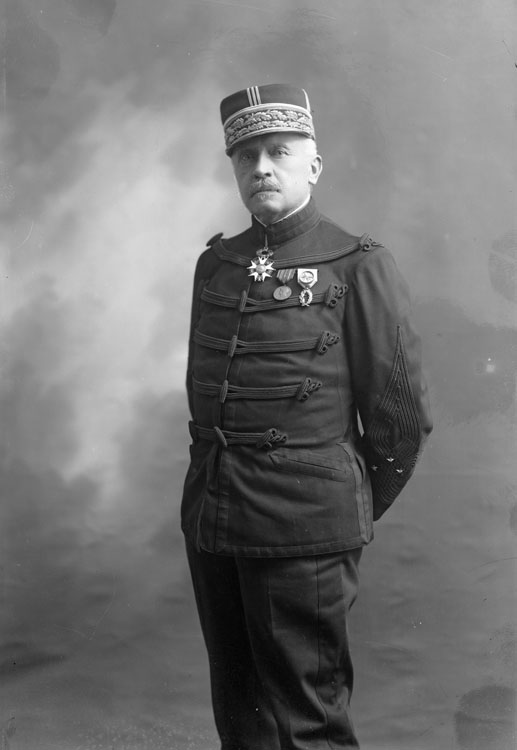 Nicolas Charles Chomer, en uniforme militaire de général de division en petite tenue de la Troisième République. En mi-corps, debout, pose de face, portant la médaille de la Légion d'Honneur et son képi sur la tête, les mains derrière son dos.