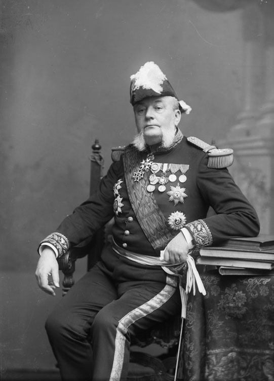 Armand Besnard, vice-amiral et ministre de la Marine de 1895 à 1898, portant l'uniforme militaire. En mi-corps posant assis sur un fauteuil, de face, devant une toile de fond représentant une base de pilier, un bras accoudé sur des livres se trouvant sur un coin de table.