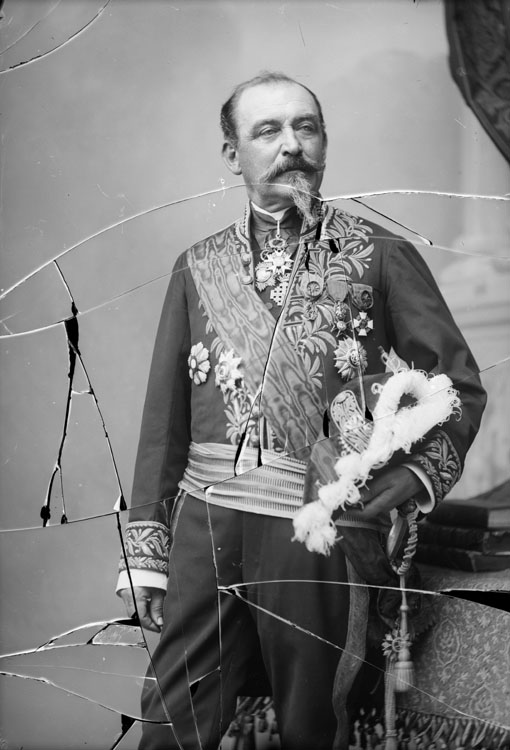 Tiburce Féraud en costume de préfet, tenant son bicorne sur le bras. En mi-corps, pose de trois quarts, à coté d'une table.