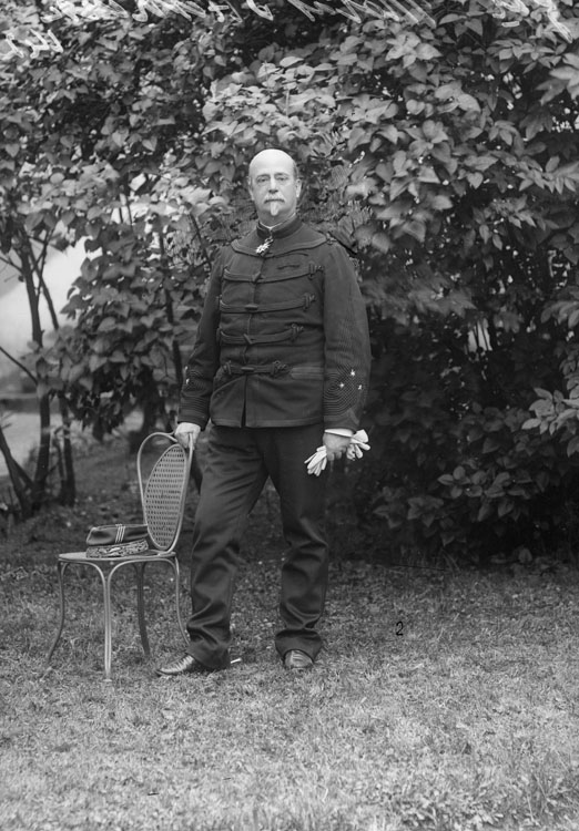 Eugène Motas-d’Hestreux, en uniforme militaire de général de brigade, posant en extérieur devant des feuillages. En pied, pose de face, une main posée sur le dossier d'une chaise et de l'autre tenant ses gants, képi sur la chaise.